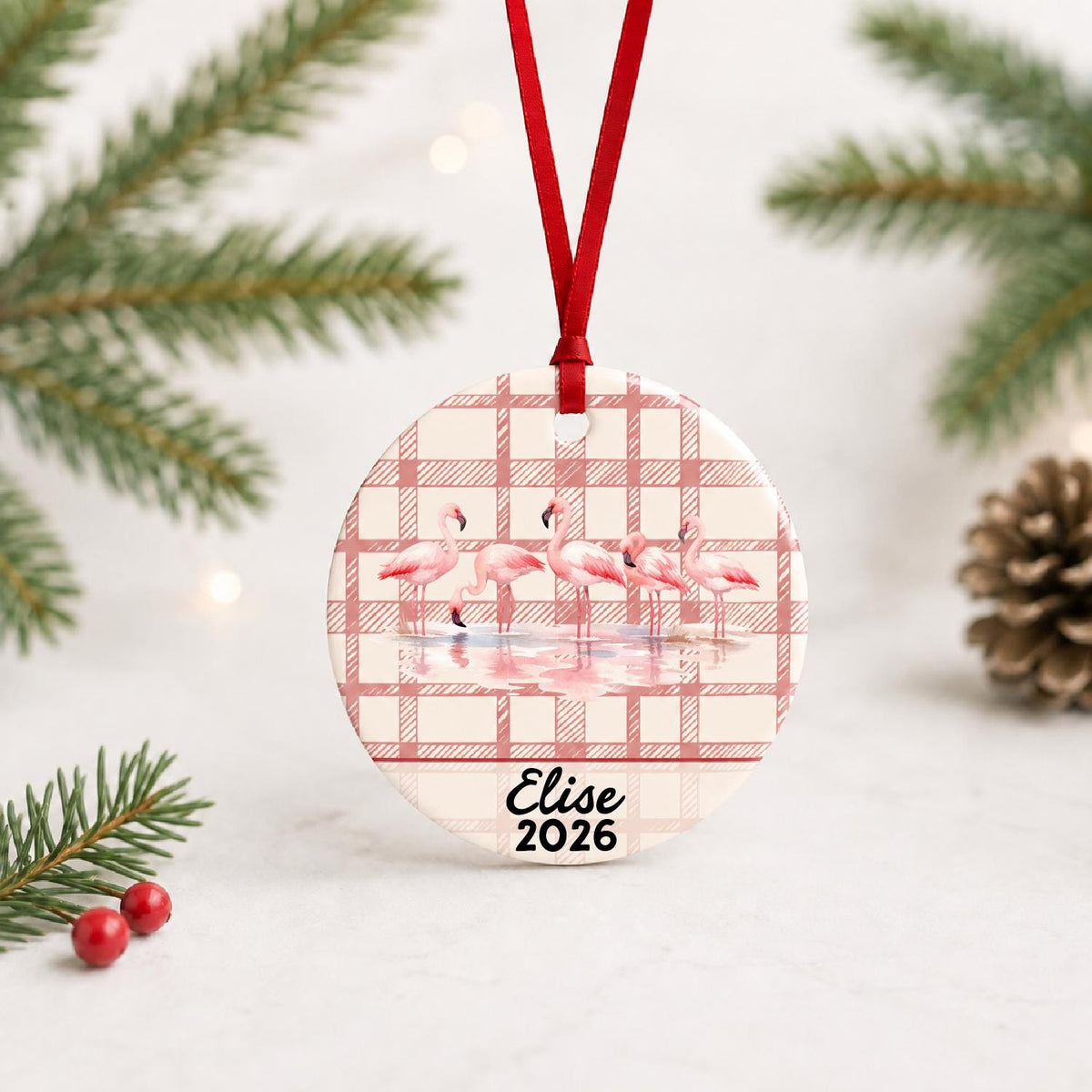Personalized flamingo flock ceramic ornament with name and year on red plaid with red ribbon on a white surface with pine branches and pinecones