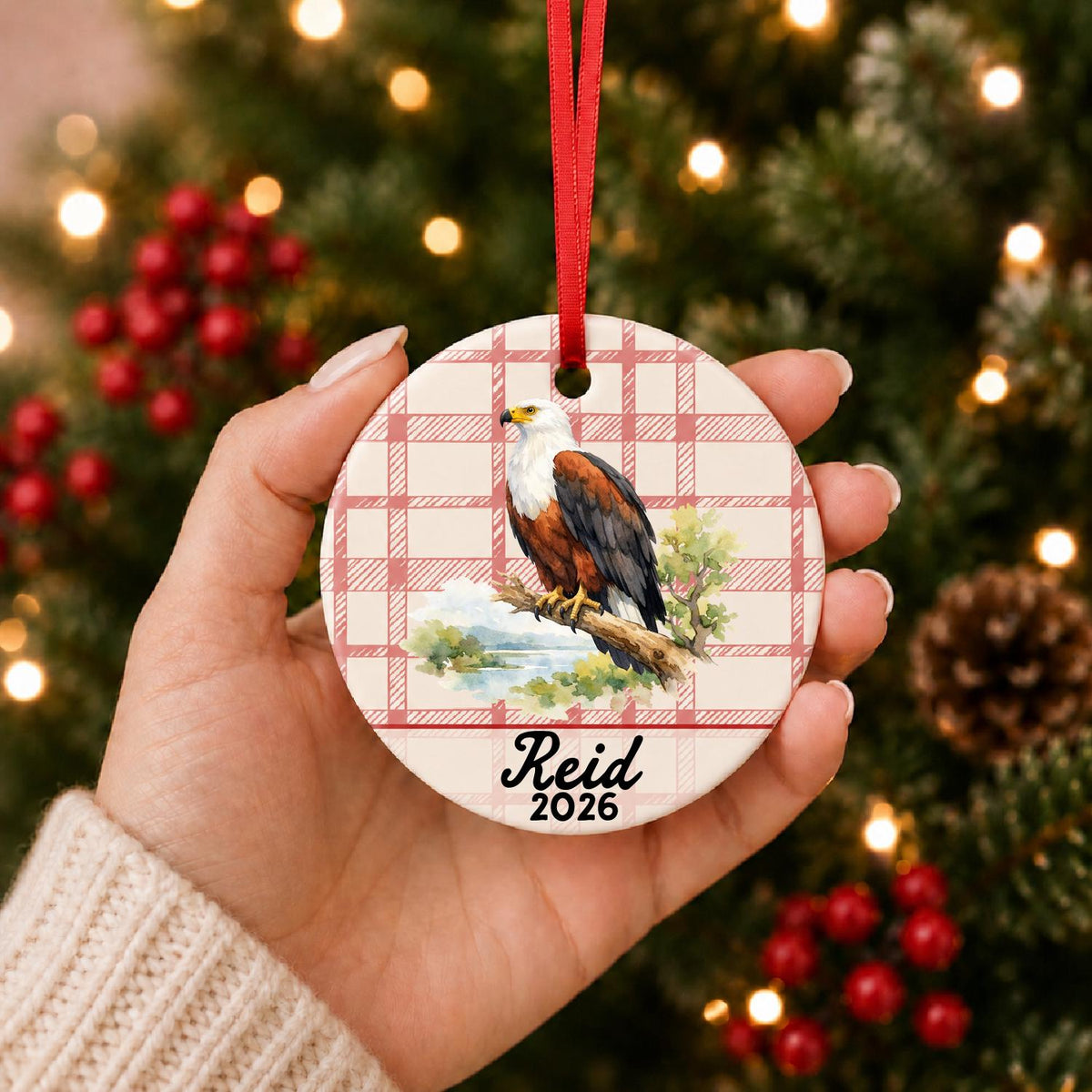 Hand holding the personalized african fish eagle ceramic ornament with name and year on red plaid in front of a Christmas tree