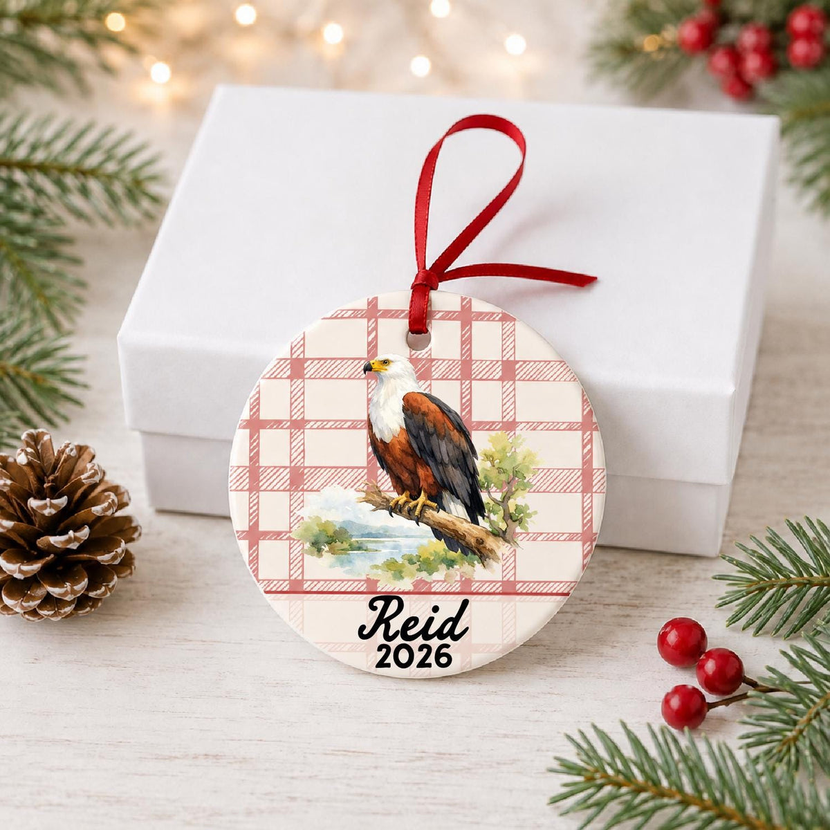 Personalized african fish eagle ceramic ornament with name and year on red plaid in front of a white gift box with pinecones and red berries