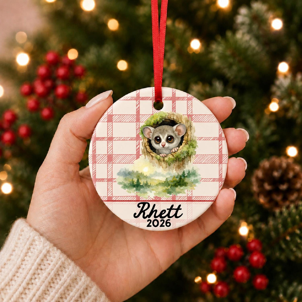 Hand holding the personalized galago ceramic ornament with name and year on red plaid in front of a Christmas tree