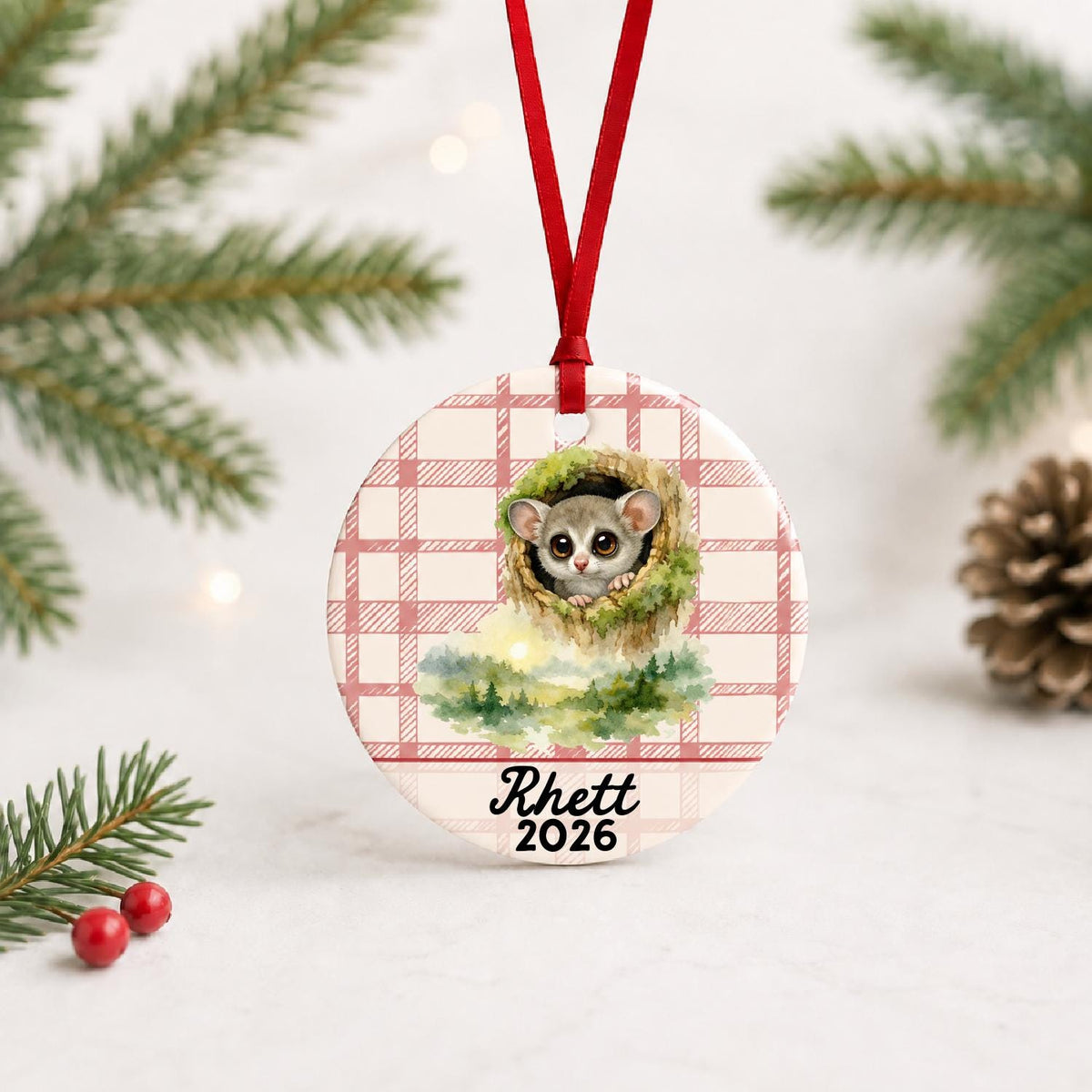 Personalized galago ceramic ornament with name and year on red plaid with red ribbon on a white surface with pine branches and pinecones
