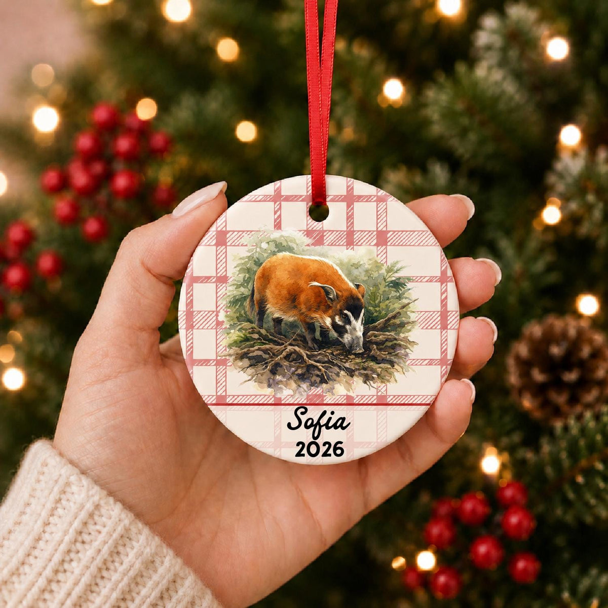 Hand holding the personalized red river hog in forest ceramic ornament with name and year on red plaid in front of a Christmas tree