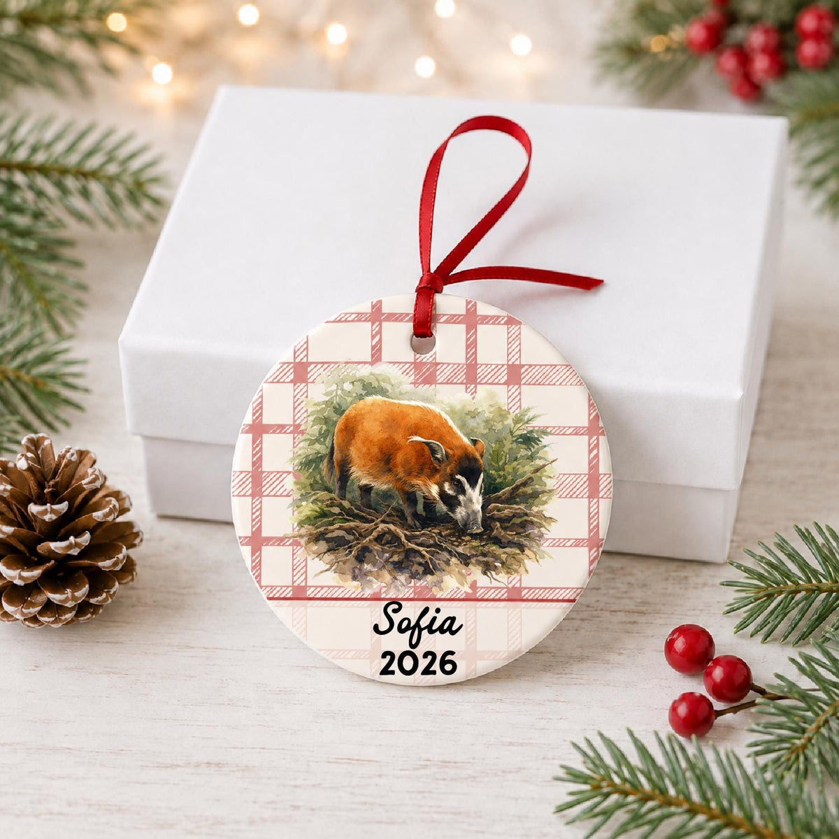 Personalized red river hog in forest ceramic ornament with name and year on red plaid in front of a white gift box with pinecones and red berries
