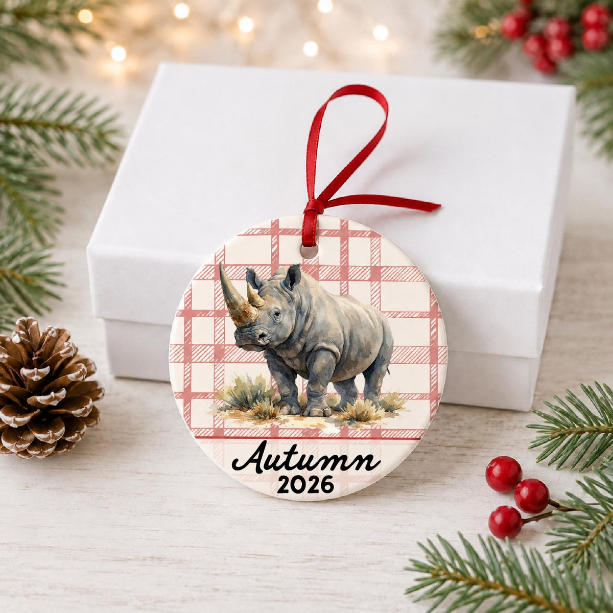 Personalized white rhinoceros in grassland ceramic ornament with name and year on red plaid in front of a white gift box with pinecones and red berries