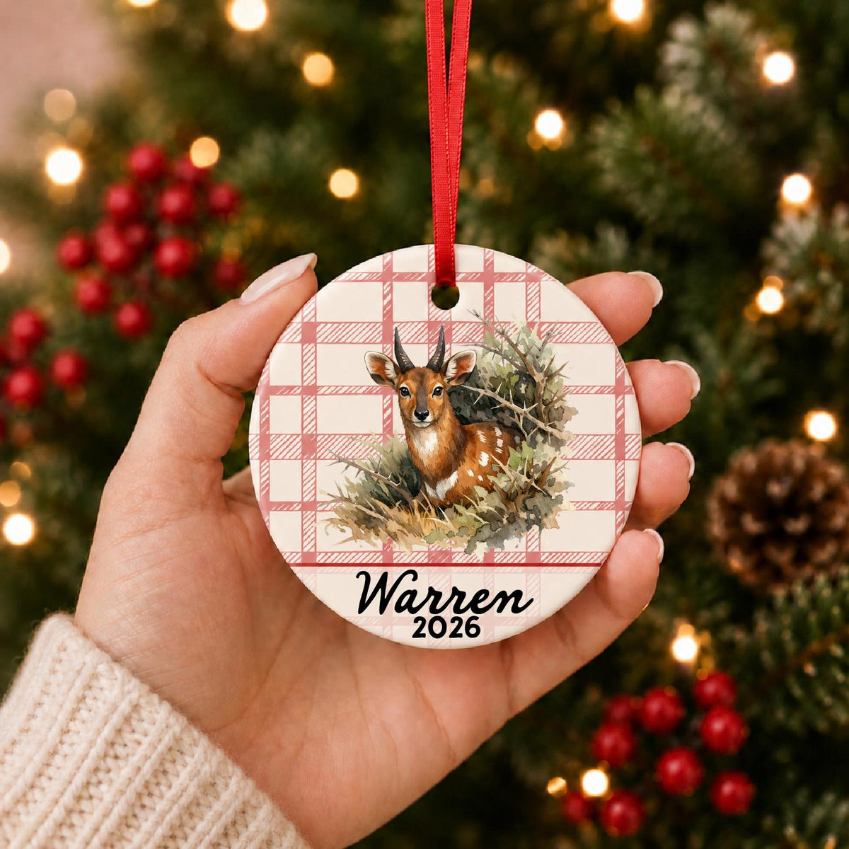 Hand holding the personalized bushbuck ceramic ornament with name and year on red plaid in front of a Christmas tree