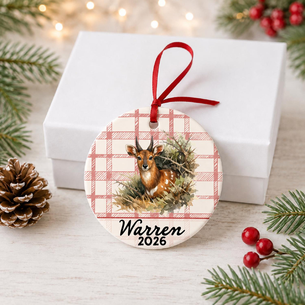 Personalized bushbuck ceramic ornament with name and year on red plaid in front of a white gift box with pinecones and red berries