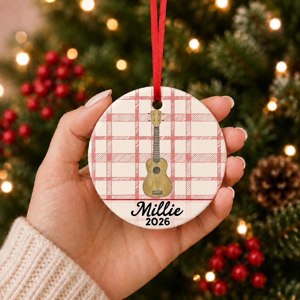 Hand holding the personalized ukulele ceramic ornament with name and year on red plaid in front of a Christmas tree