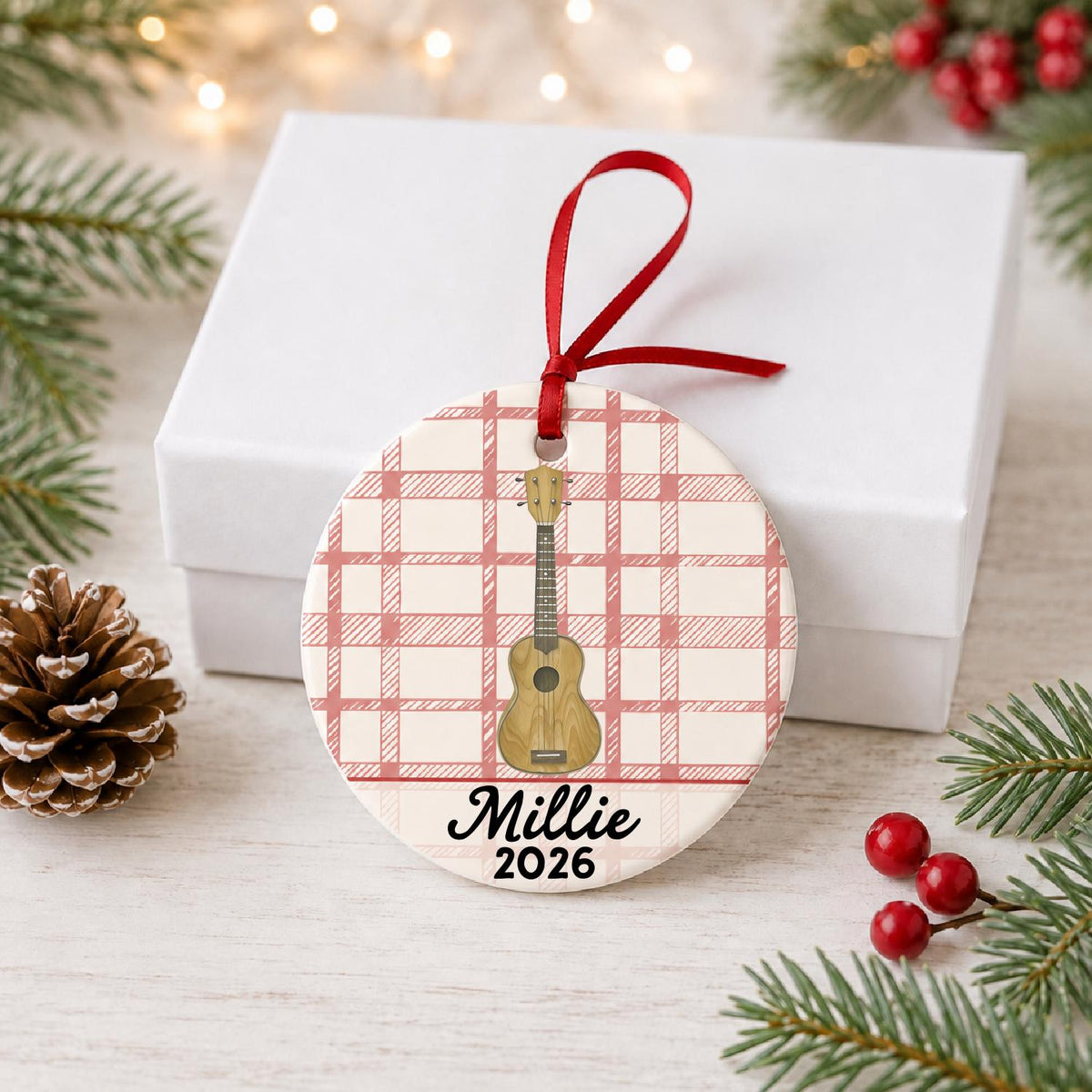 Personalized ukulele ceramic ornament with name and year on red plaid in front of a white gift box with pinecones and red berries
