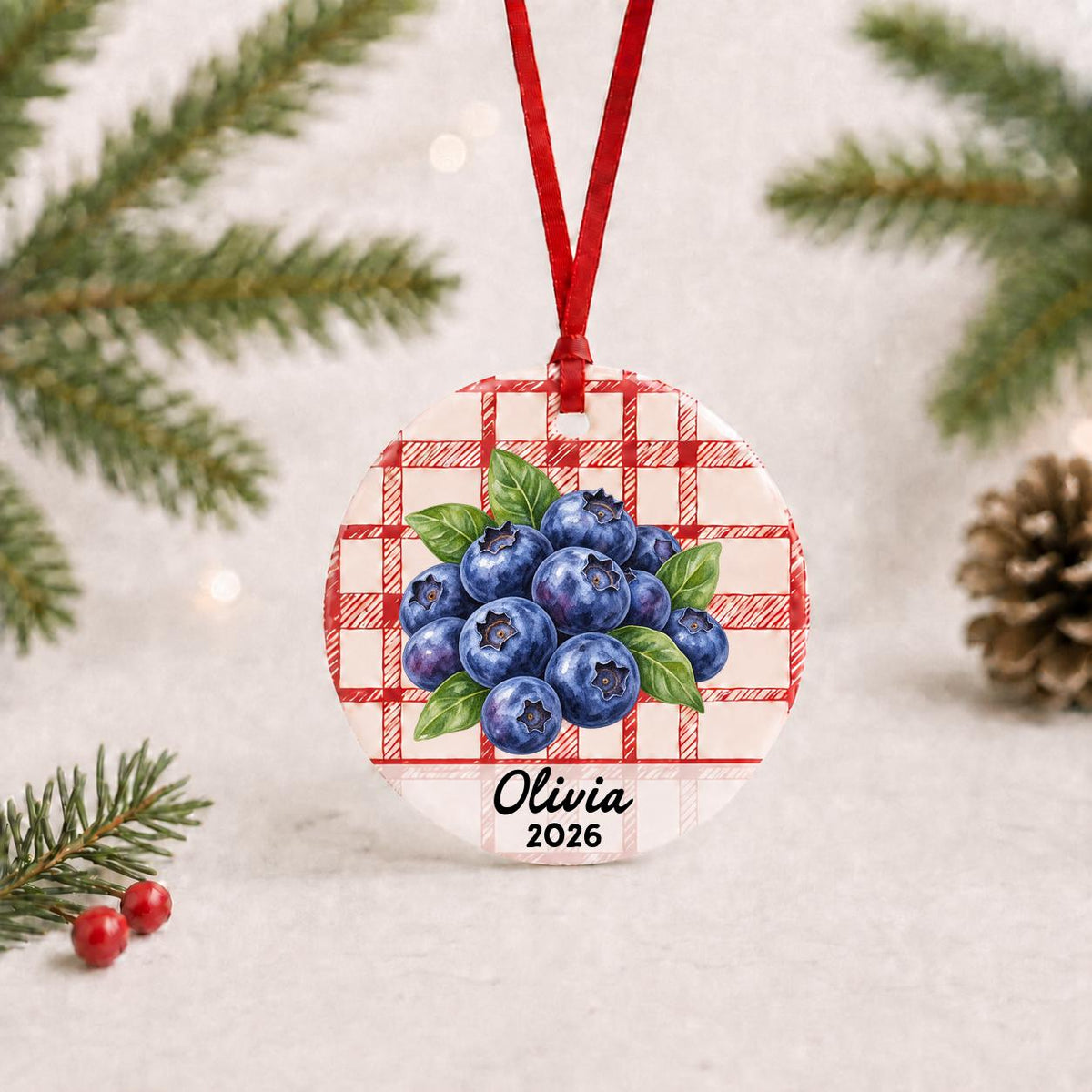 Personalized blueberries ceramic ornament with name and year on red plaid with red ribbon on a white surface with pine branches and pinecones