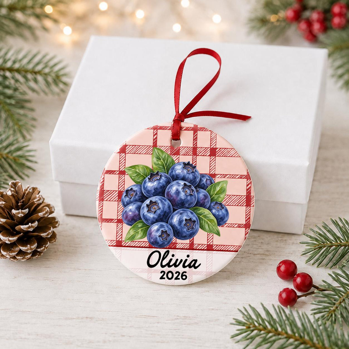 Personalized blueberries ceramic ornament with name and year on red plaid in front of a white gift box with pinecones and red berries