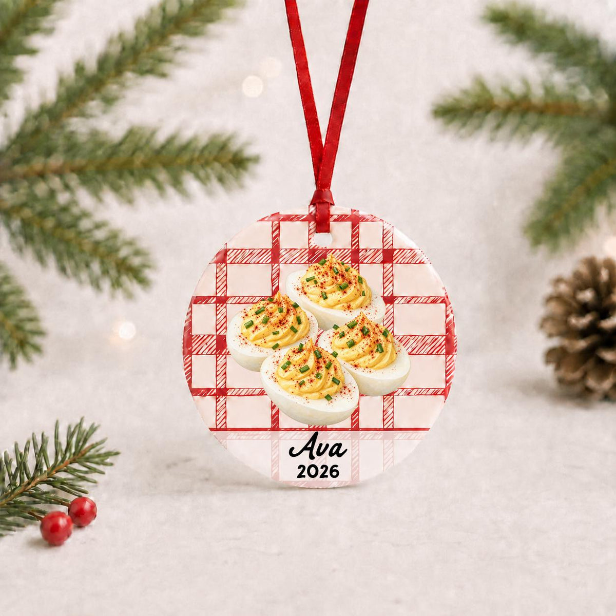 Personalized deviled eggs ceramic ornament with name and year on red plaid with red ribbon on a white surface with pine branches and pinecones