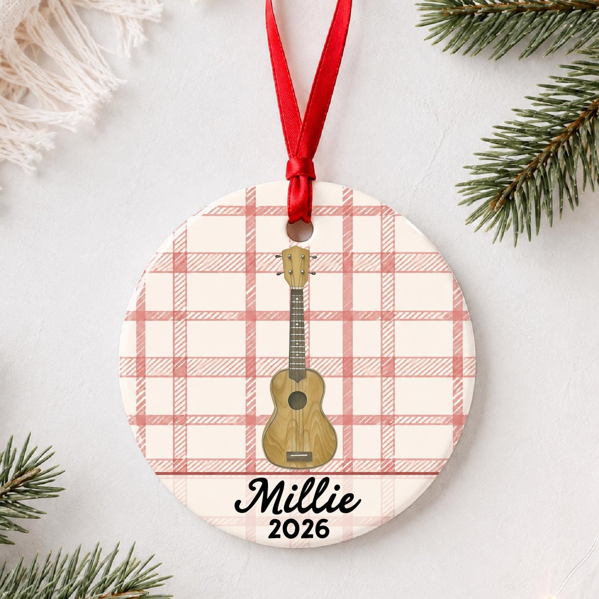 Personalized ukulele ceramic ornament with name and year on red plaid styled with pine branches on a soft white background