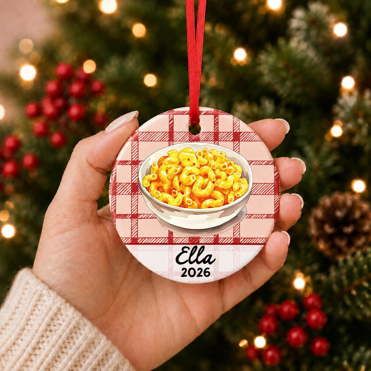 Hand holding the personalized mac and cheese ceramic ornament with name and year on red plaid in front of a Christmas tree