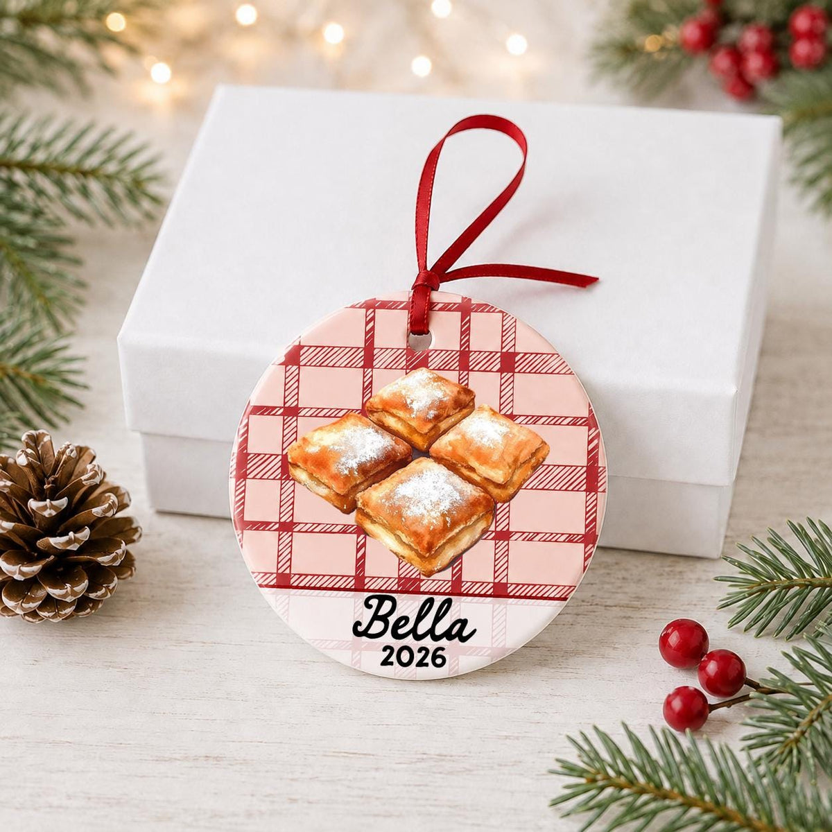 Personalized beignets ceramic ornament with name and year on red plaid in front of a white gift box with pinecones and red berries