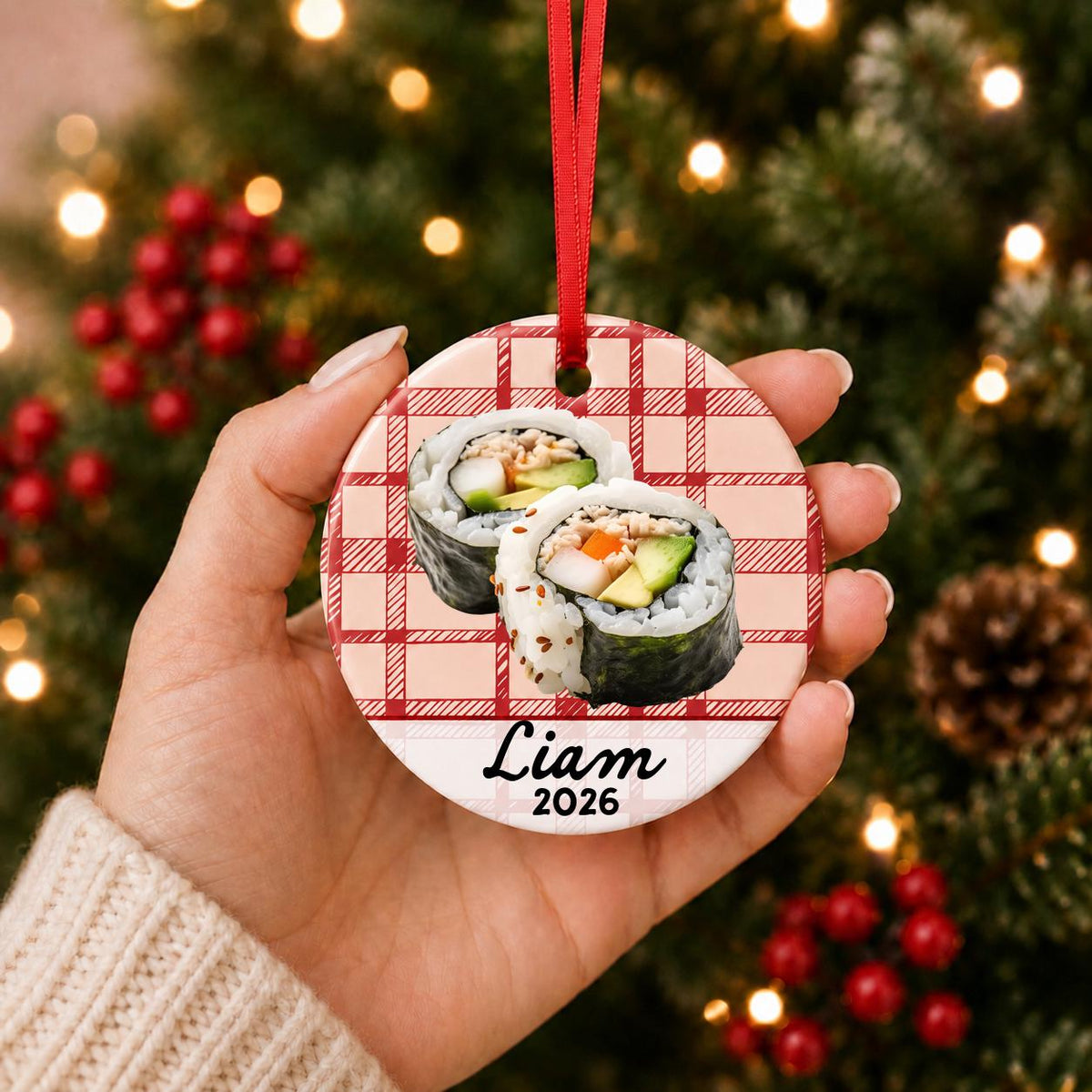 Hand holding the personalized sushi ceramic ornament with name and year on red plaid in front of a Christmas tree