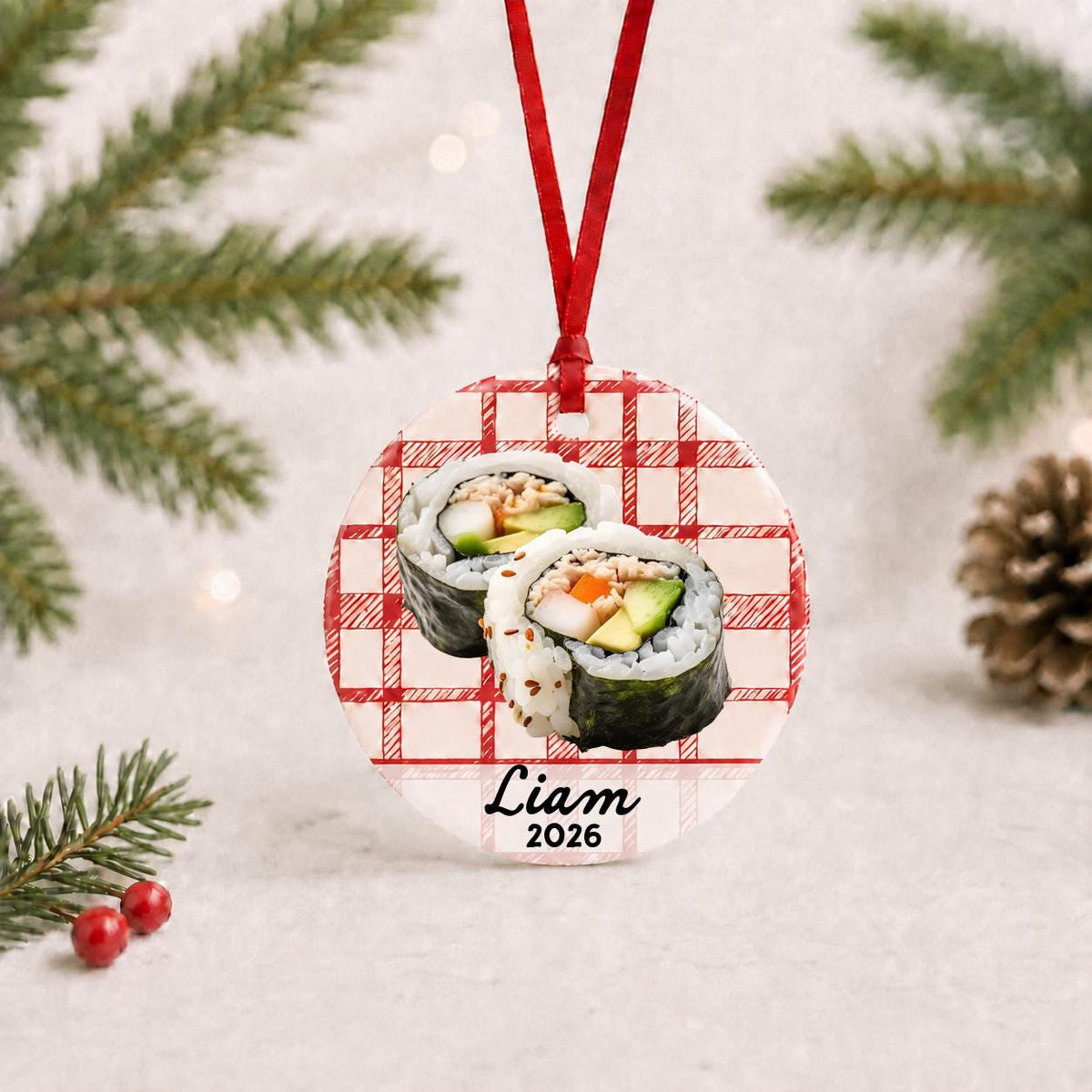 Personalized sushi ceramic ornament with name and year on red plaid with red ribbon on a white surface with pine branches and pinecones