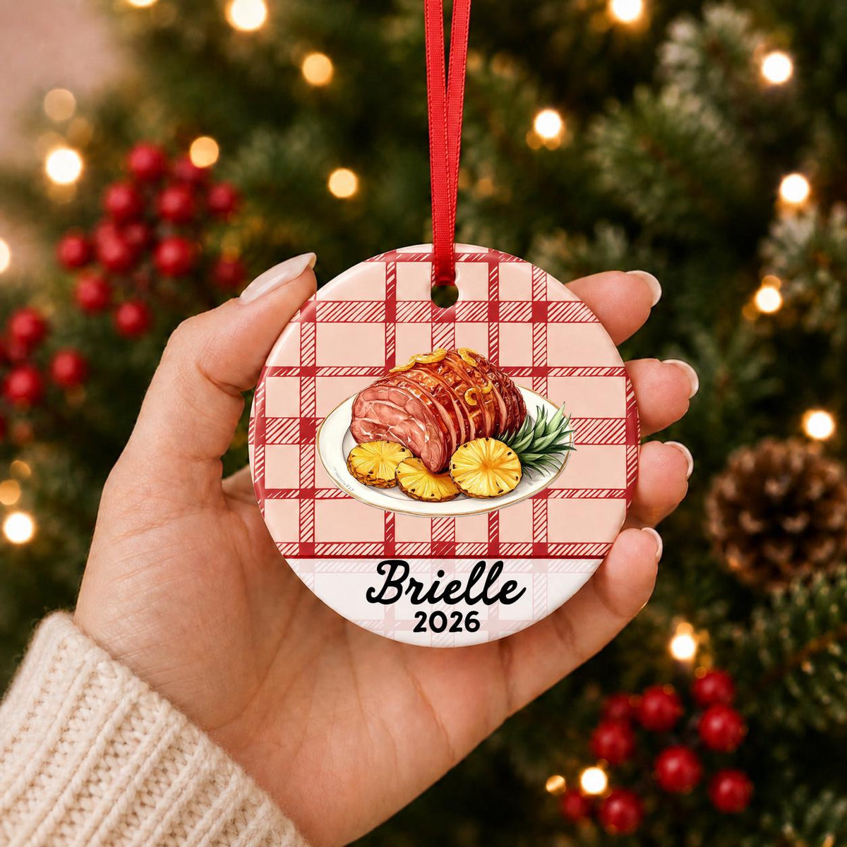 Hand holding the personalized Christmas ham ceramic ornament with name and year on red plaid in front of a Christmas tree