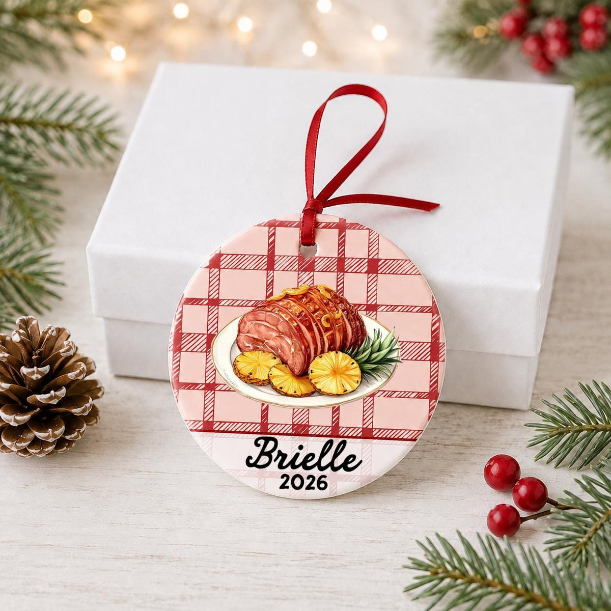 Personalized Christmas ham ceramic ornament with name and year on red plaid in front of a white gift box with pinecones and red berries