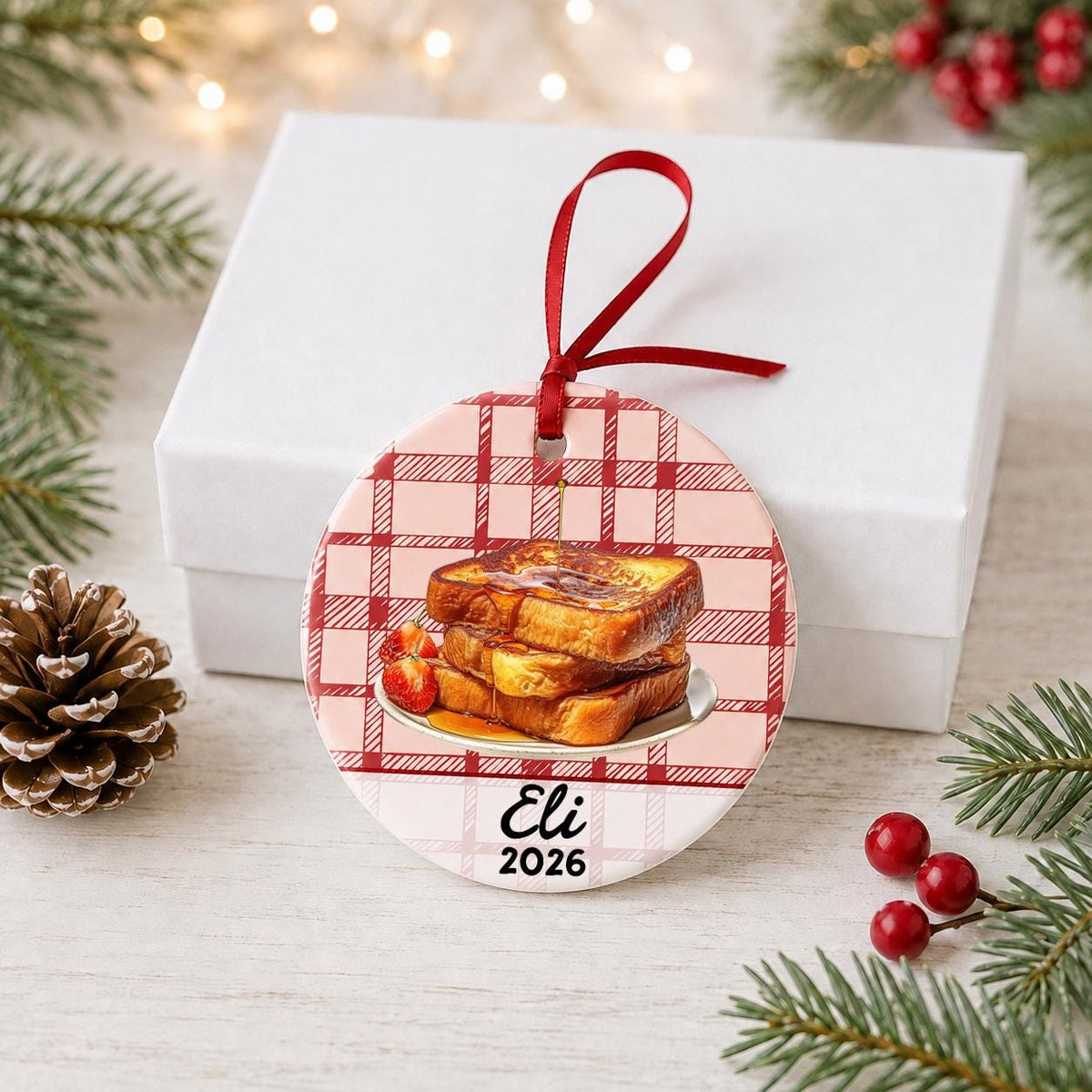 Personalized French toast ceramic ornament with name and year on red plaid in front of a white gift box with pinecones and red berries