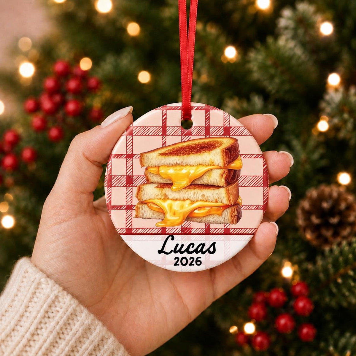 Hand holding the personalized grilled cheese ceramic ornament with name and year on red plaid in front of a Christmas tree