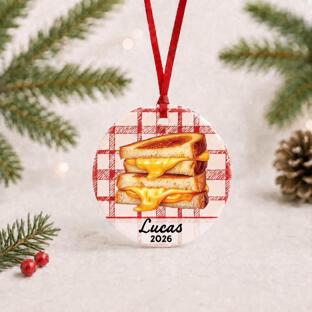 Personalized grilled cheese ceramic ornament with name and year on red plaid with red ribbon on a white surface with pine branches and pinecones