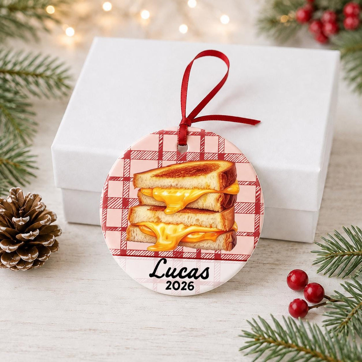 Personalized grilled cheese ceramic ornament with name and year on red plaid in front of a white gift box with pinecones and red berries