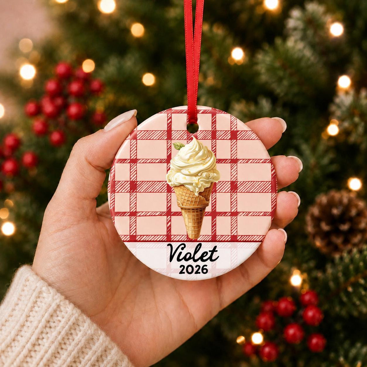 Hand holding the personalized soft serve ice cream ceramic ornament with name and year on red plaid in front of a Christmas tree