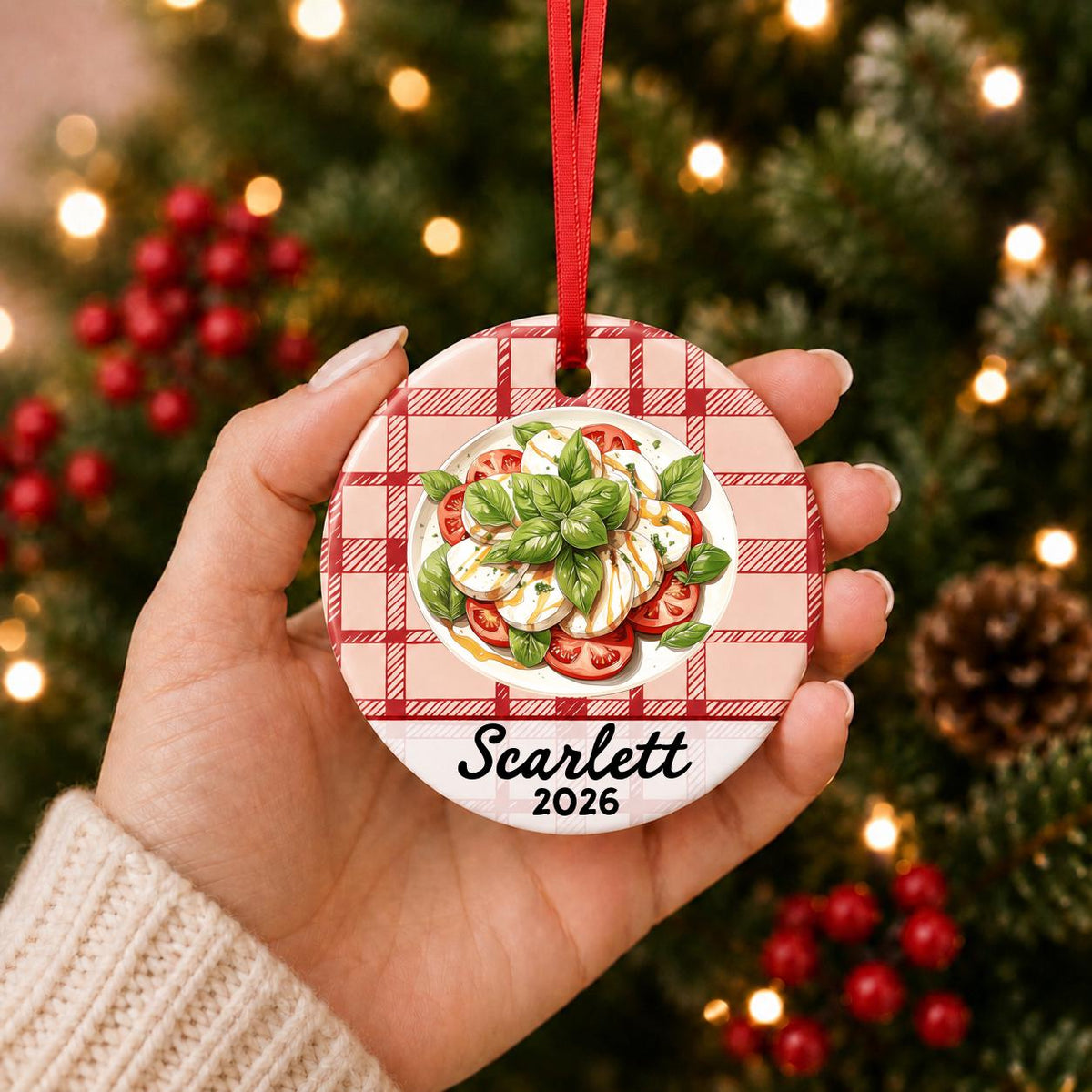 Hand holding the personalized caprese salad ceramic ornament with name and year on red plaid in front of a Christmas tree