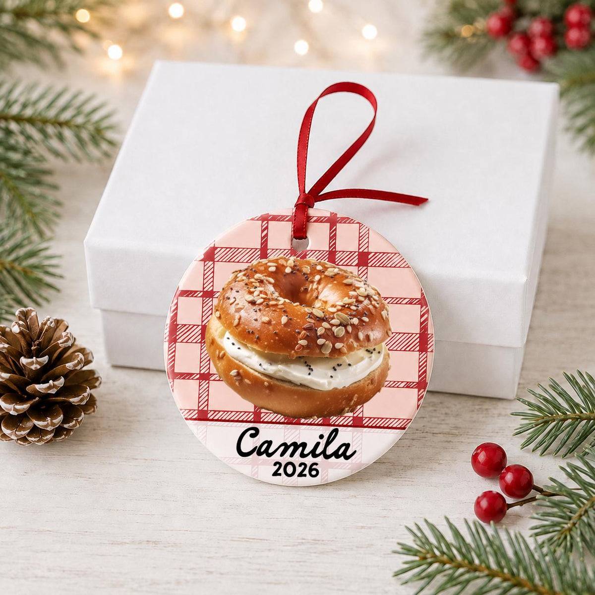 Personalized bagel ceramic ornament with name and year on red plaid in front of a white gift box with pinecones and red berries