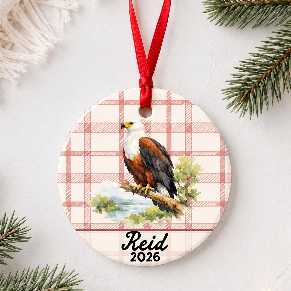 Personalized african fish eagle ceramic ornament with name and year on red plaid styled with pine branches on a soft white background