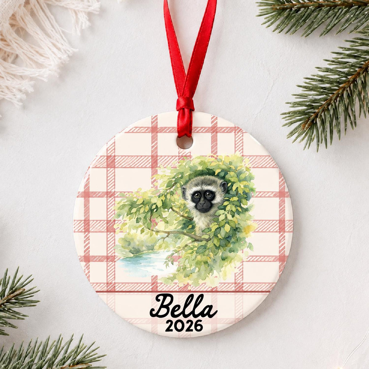 Personalized vervet monkey ceramic ornament with name and year on red plaid styled with pine branches on a soft white background