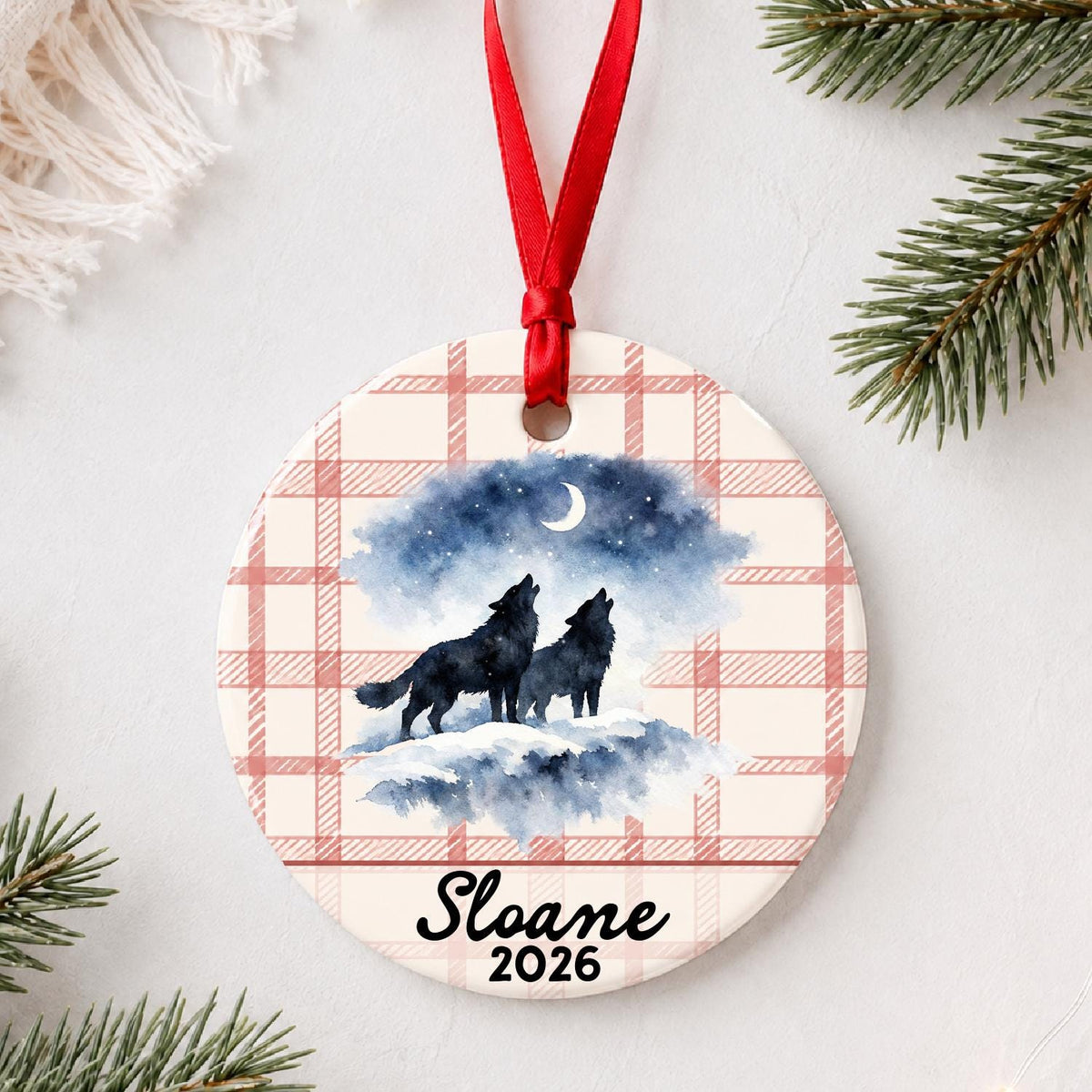 Personalized howling wolf pair ceramic ornament with name and year on red plaid styled with pine branches on a soft white background
