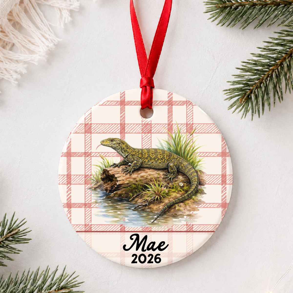 Personalized monitor lizard ceramic ornament with name and year on red plaid styled with pine branches on a soft white background