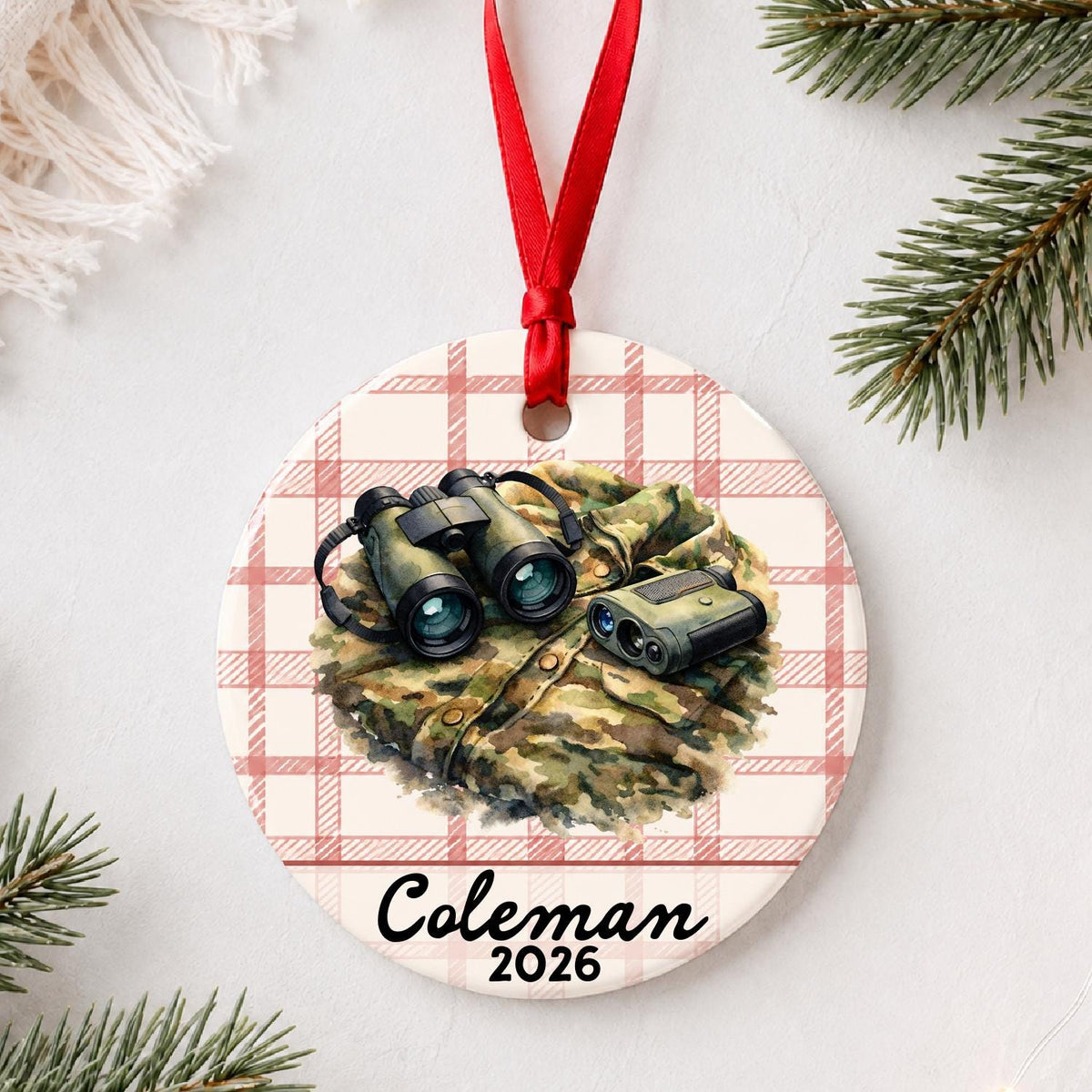 Personalized binoculars and rangefinder ceramic ornament with name and year on red plaid styled with pine branches on a soft white background