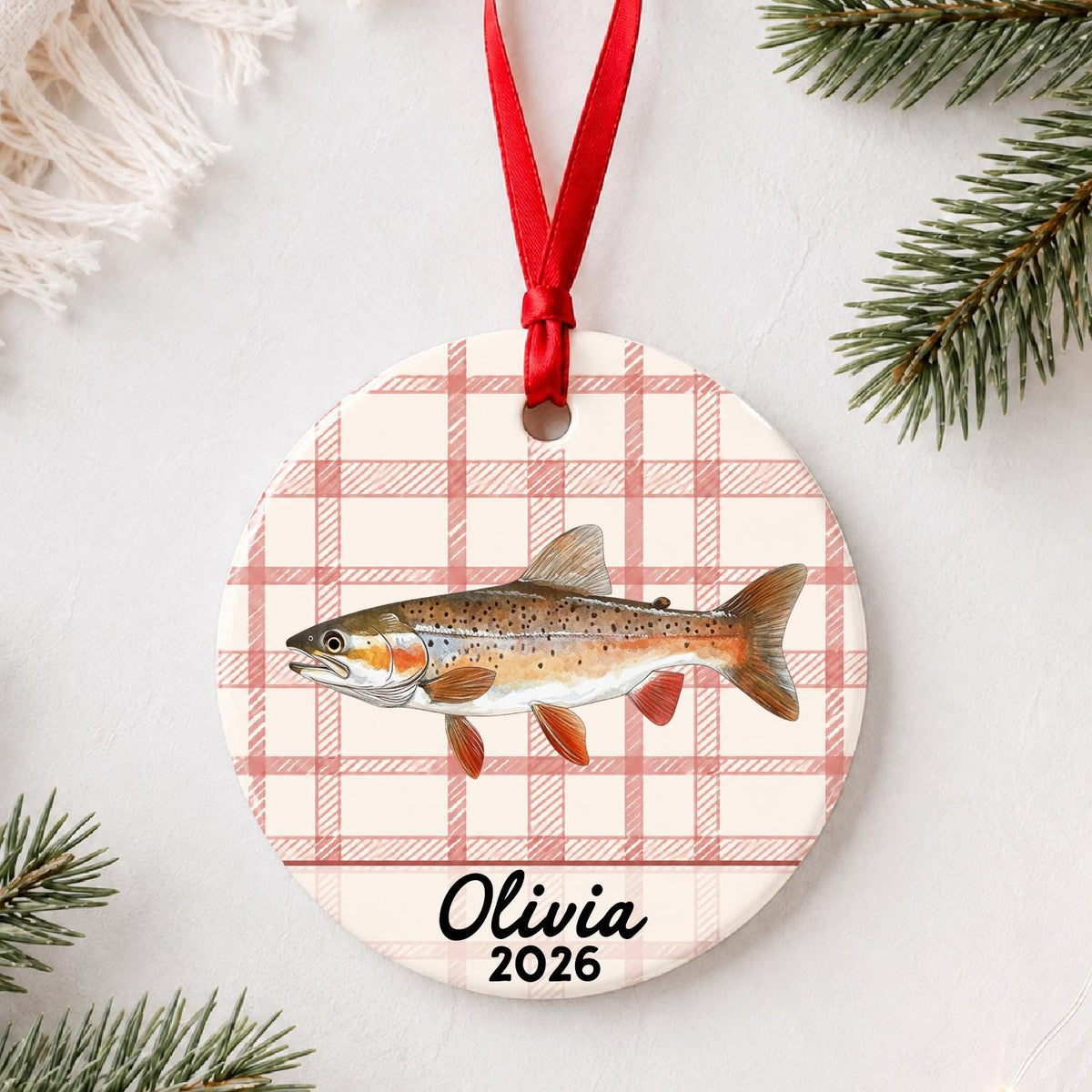 Personalized rainbow trout ceramic ornament with name and year on red plaid styled with pine branches on a soft white background