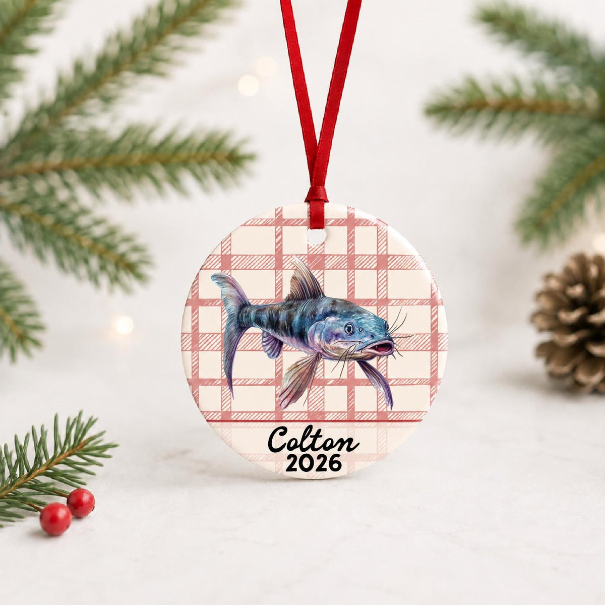 Personalized catfish ceramic ornament with name and year on red plaid with red ribbon on a white surface with pine branches and pinecones