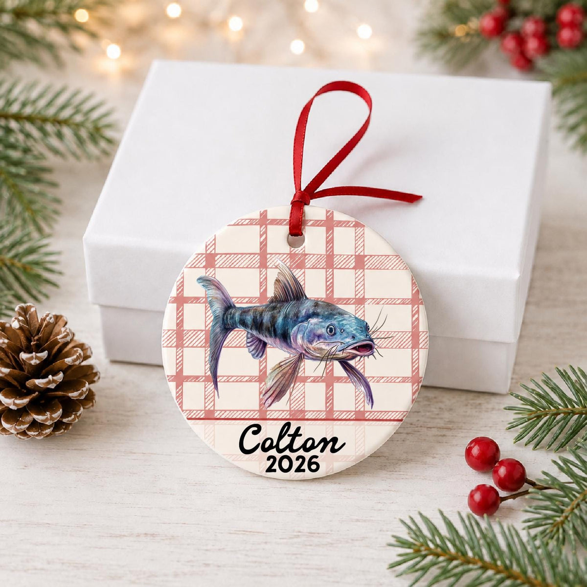Personalized catfish ceramic ornament with name and year on red plaid in front of a white gift box with pinecones and red berries
