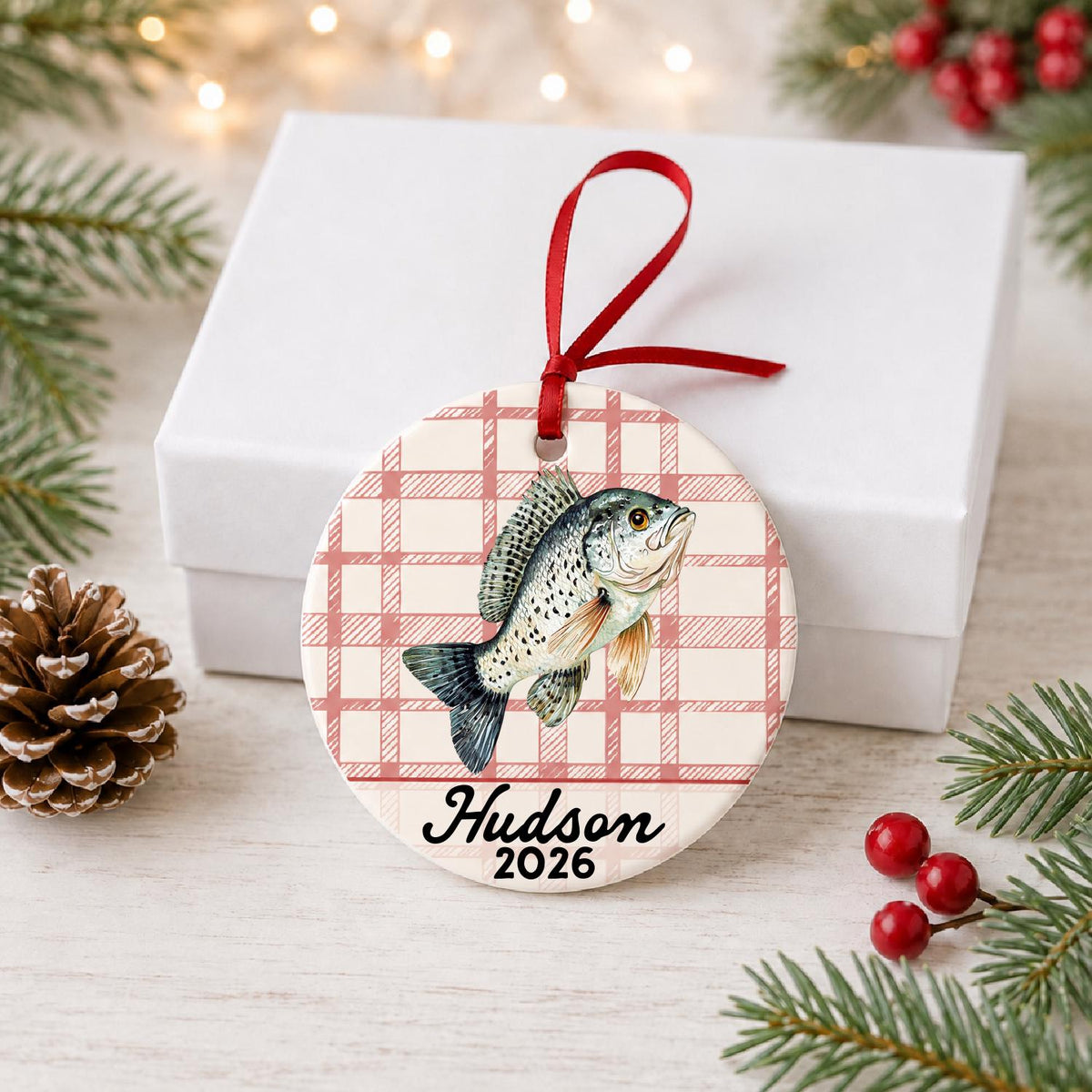 Personalized crappie ceramic ornament with name and year on red plaid in front of a white gift box with pinecones and red berries