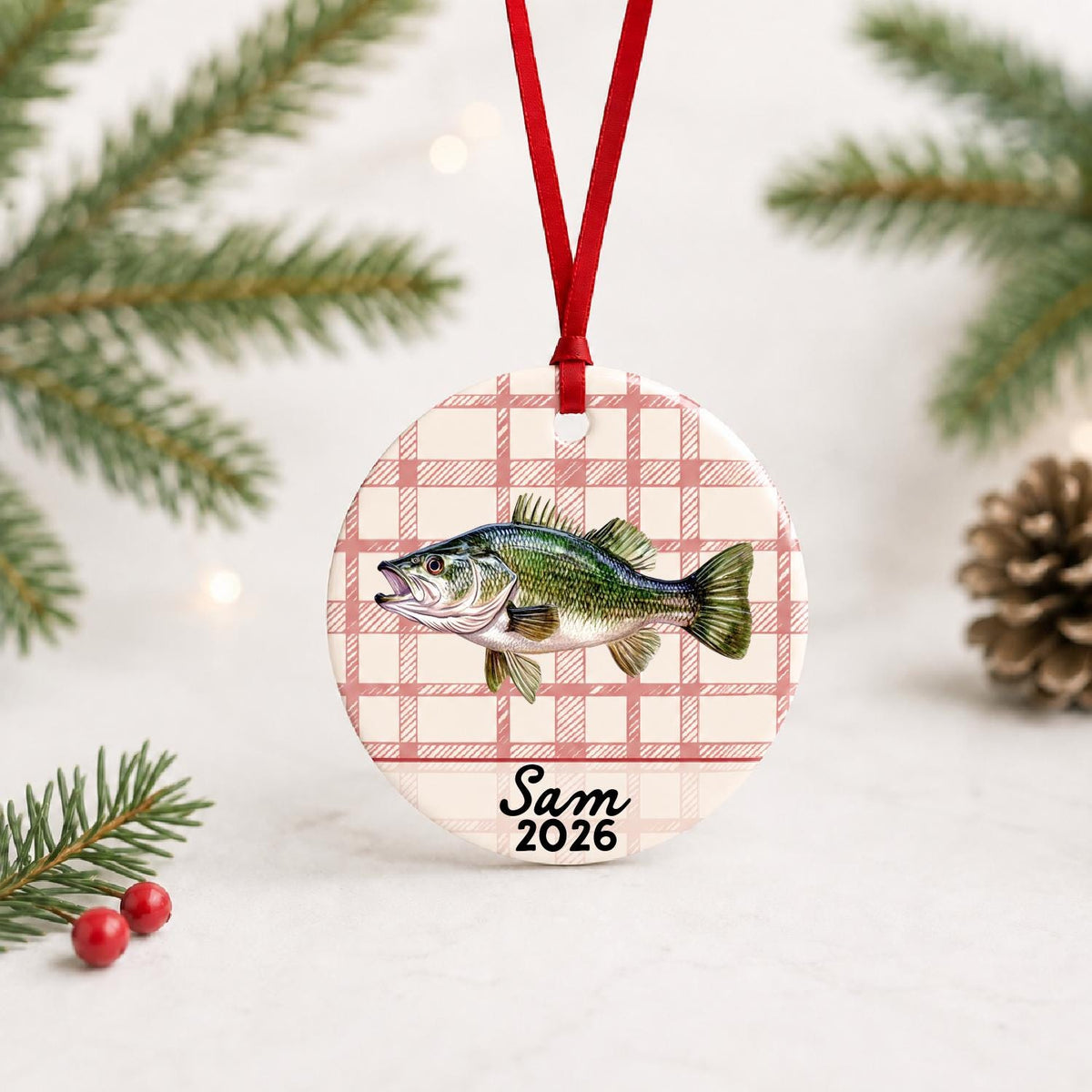 Personalized largemouth bass ceramic ornament with name and year on red plaid with red ribbon on a white surface with pine branches and pinecones