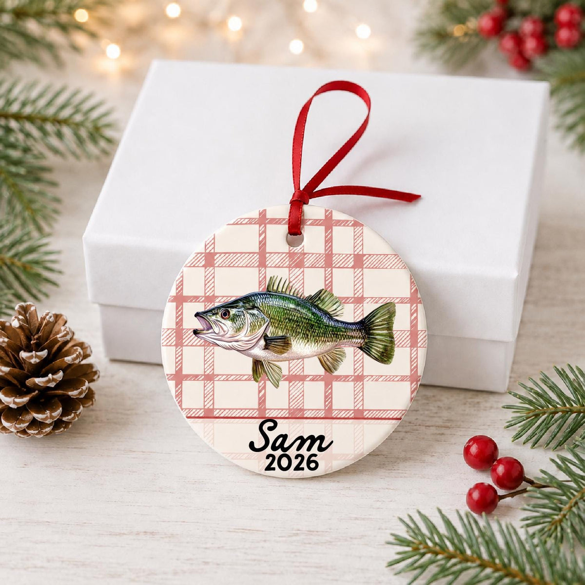 Personalized largemouth bass ceramic ornament with name and year on red plaid in front of a white gift box with pinecones and red berries