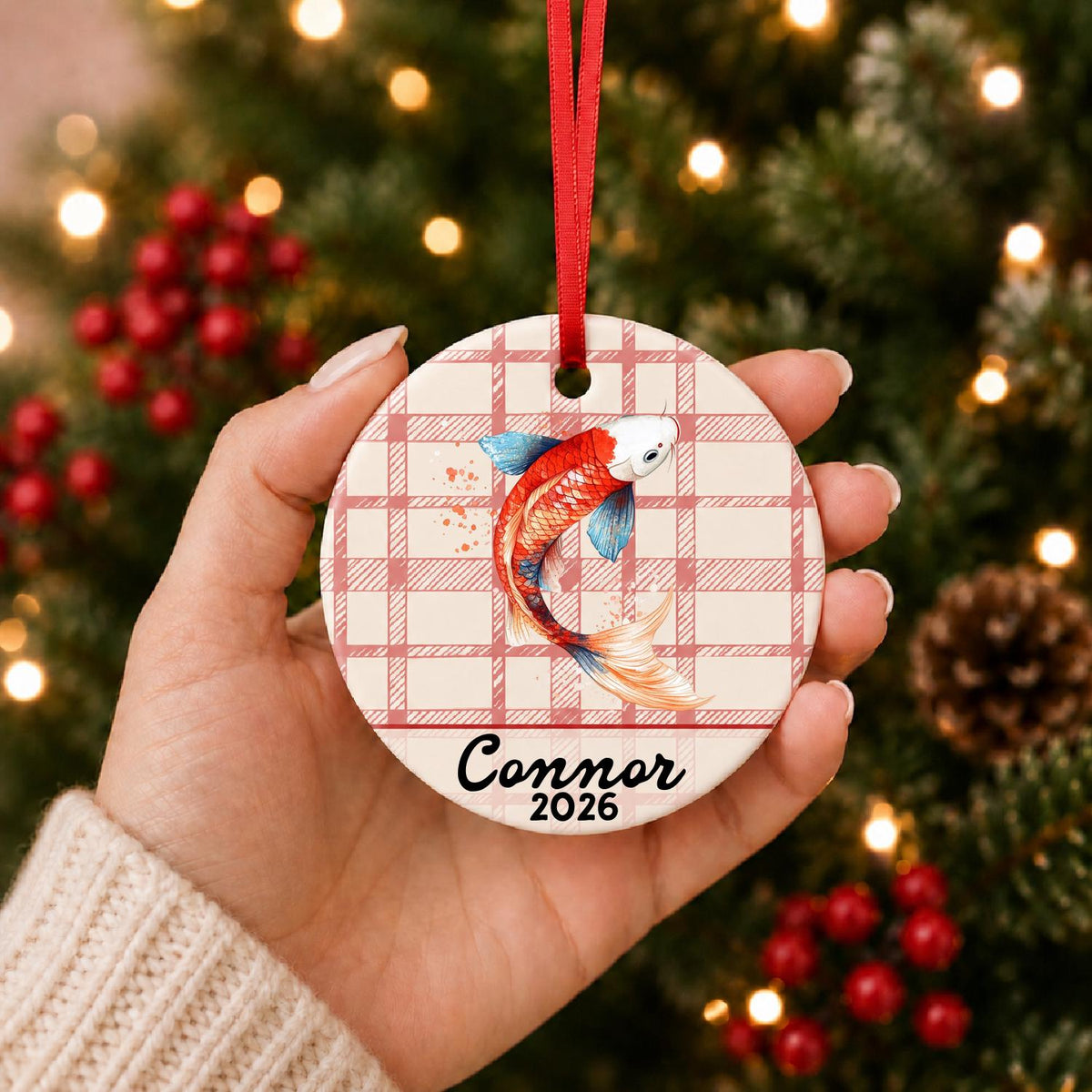 Hand holding the personalized red and white koi ceramic ornament with name and year on red plaid in front of a Christmas tree