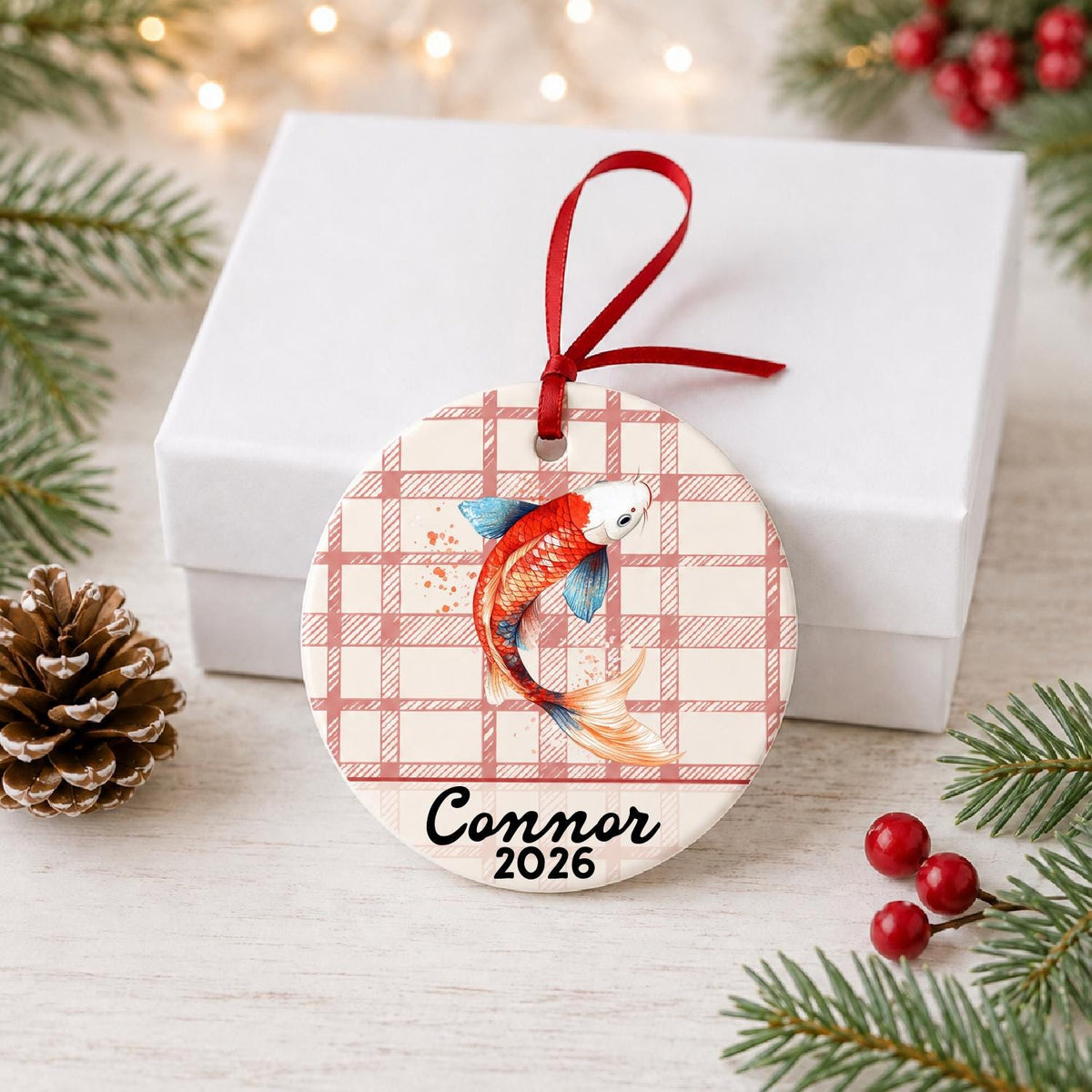 Personalized red and white koi ceramic ornament with name and year on red plaid in front of a white gift box with pinecones and red berries