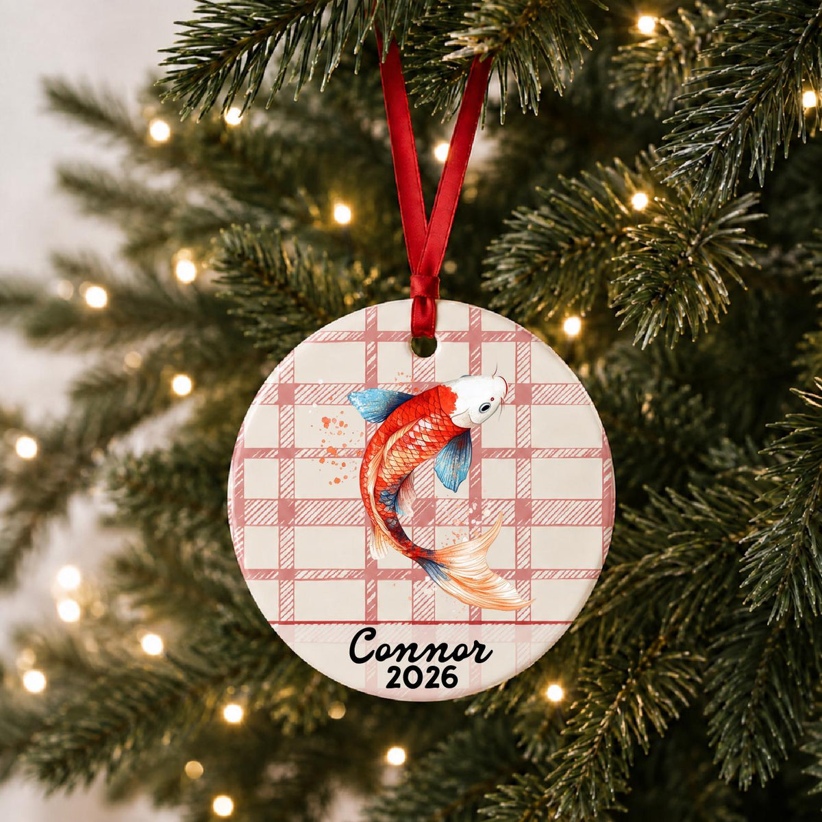 Personalized red and white koi ceramic ornament with name and year on red plaid hanging on a lit Christmas tree