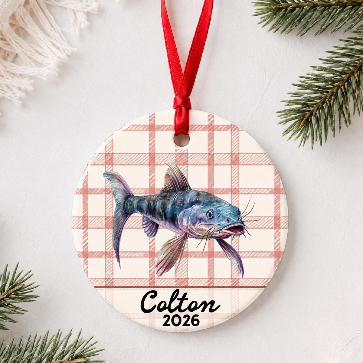 Personalized catfish ceramic ornament with name and year on red plaid styled with pine branches on a soft white background
