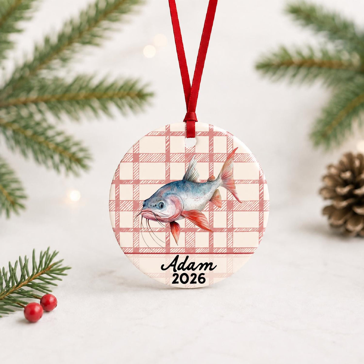 Personalized red-finned catfish ceramic ornament with name and year on red plaid with red ribbon on a white surface with pine branches and pinecones