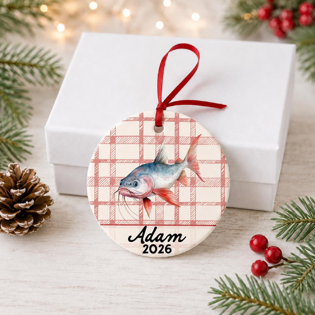 Personalized red-finned catfish ceramic ornament with name and year on red plaid in front of a white gift box with pinecones and red berries