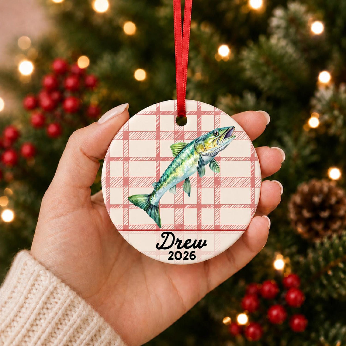 Hand holding the personalized muskie ceramic ornament with name and year on red plaid in front of a Christmas tree