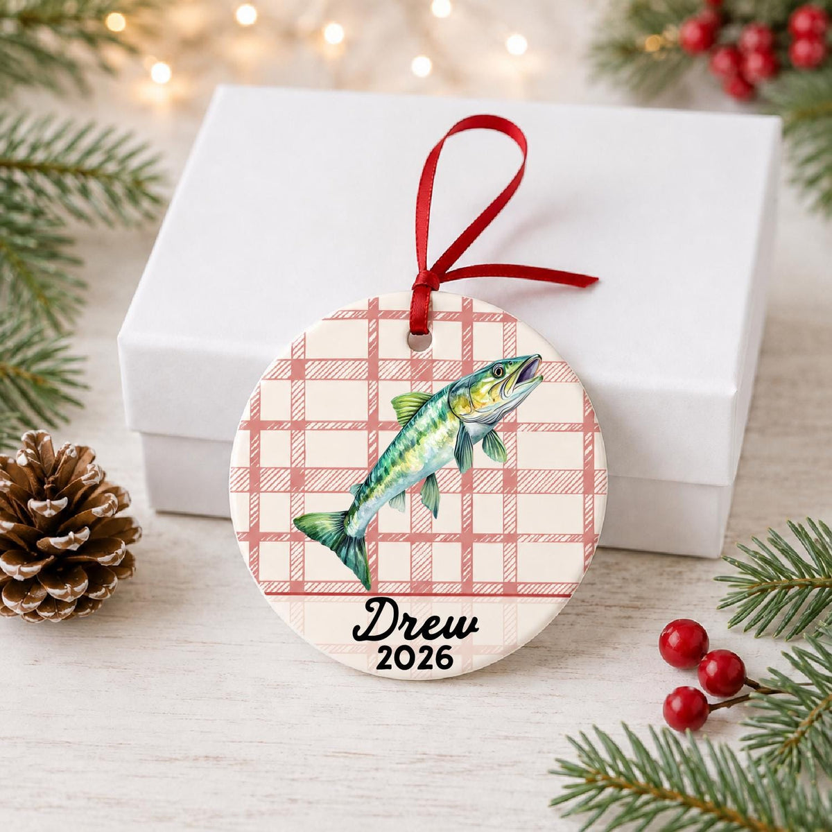Personalized muskie ceramic ornament with name and year on red plaid in front of a white gift box with pinecones and red berries