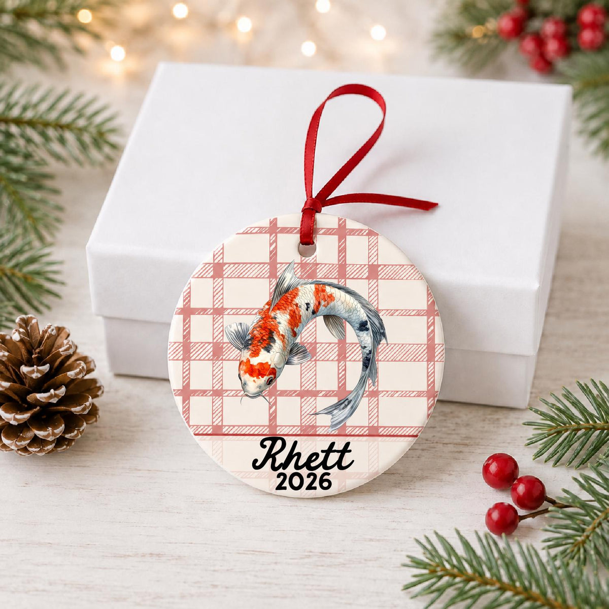 Personalized koi fish ceramic ornament with name and year on red plaid in front of a white gift box with pinecones and red berries