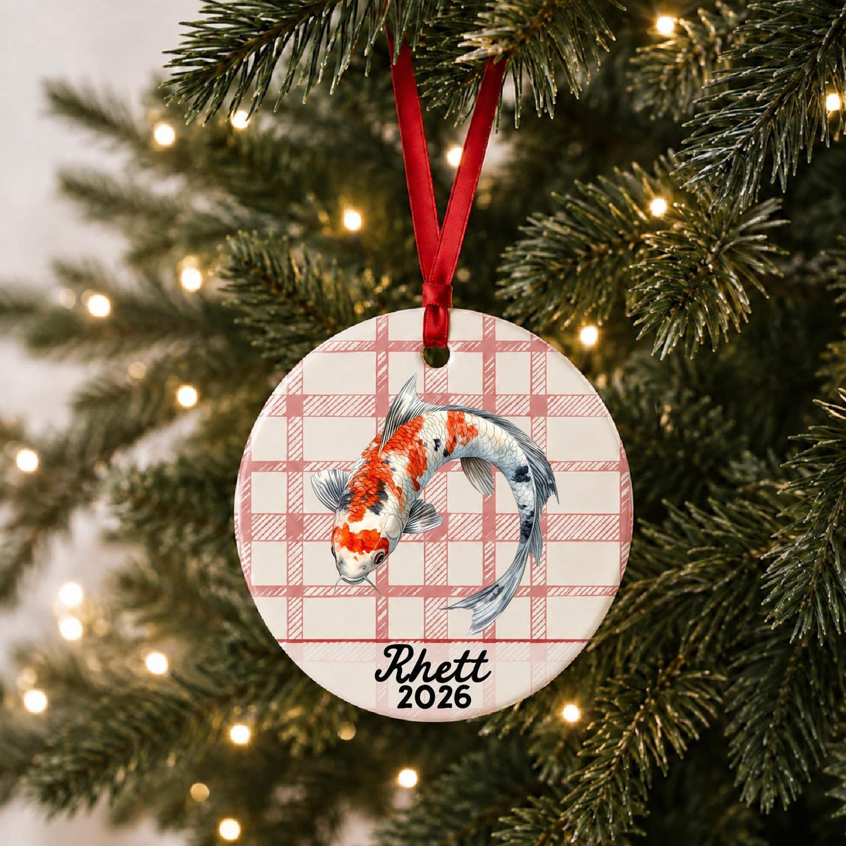 Personalized koi fish ceramic ornament with name and year on red plaid hanging on a lit Christmas tree